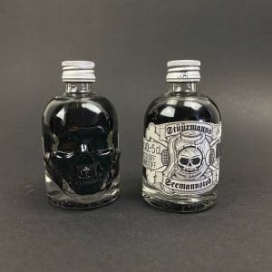 zwei Mini-Schädelflaschen mit schwarzem Stüürmanns Seemannstod Lakritz Likör, Vorderseite und Rückseite mit Etikett und Totenkopftaucher-Logo