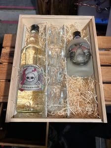 Holzbox mit durchsichtigen Stüürmanns Seemannstod Dragon Gin in der 1 Liter Gastroflasche, vier durchsichtigen Stüürmanns Schädel Schnapsgläser und blassgrüne Stüürmanns Seemannstod Dragon Gin Drachenfrucht-Gin 0,5 Liter Totenkopfflasche